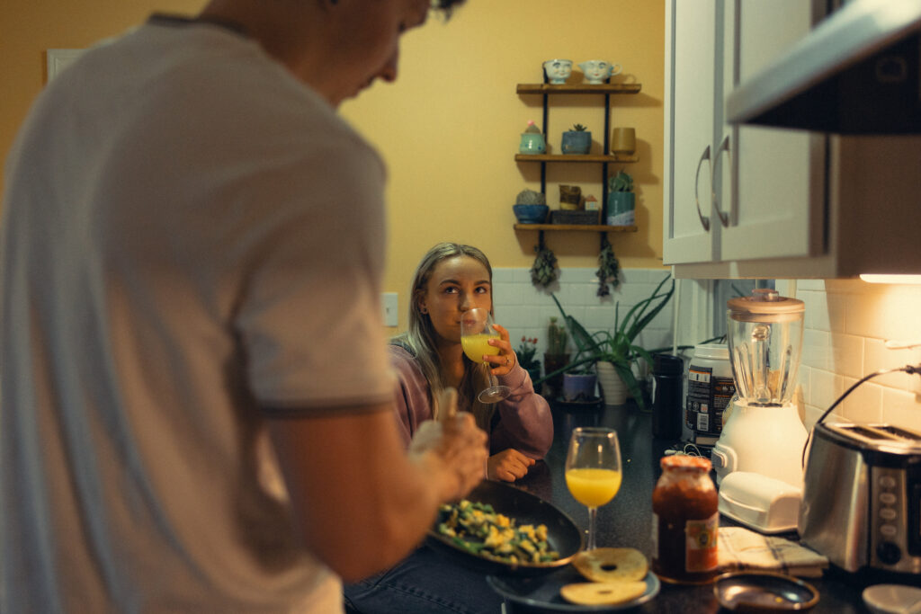 a woman sips a mimosa in the background while her fiance makes breakfast in the foreground