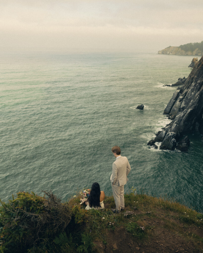 a couple sit on the edge of a cliff overlooking the ocean for their oregon elopement photography