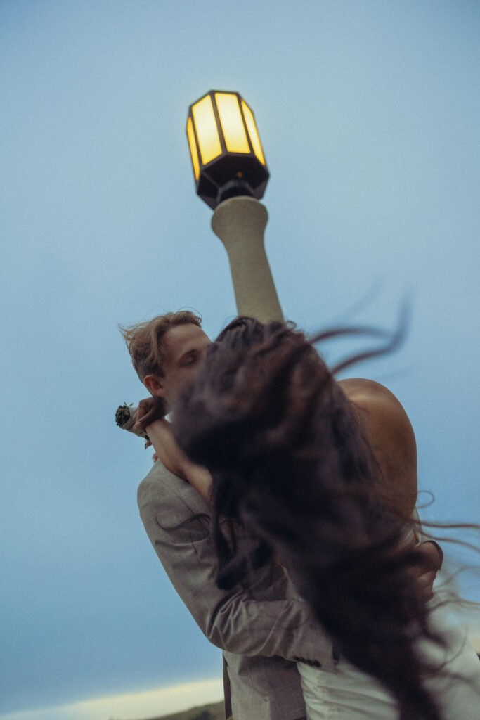 a couple stand in an embrace under a lamppost during elopement photography and her hair swishes in the wind