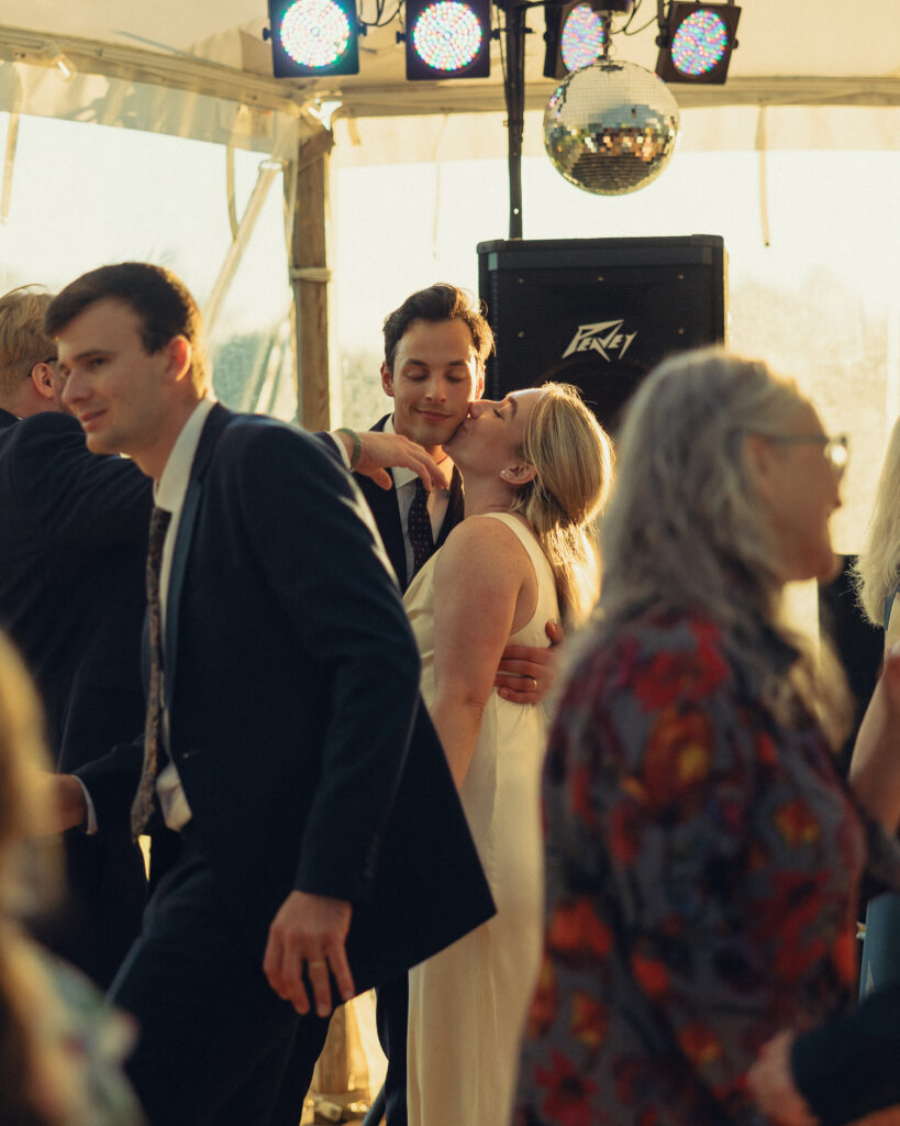 the bride presses a kiss to the grooms cheek as he holds her on the dance floor with his eyes closed surrounded by guests during the reception with documentary style wedding photos