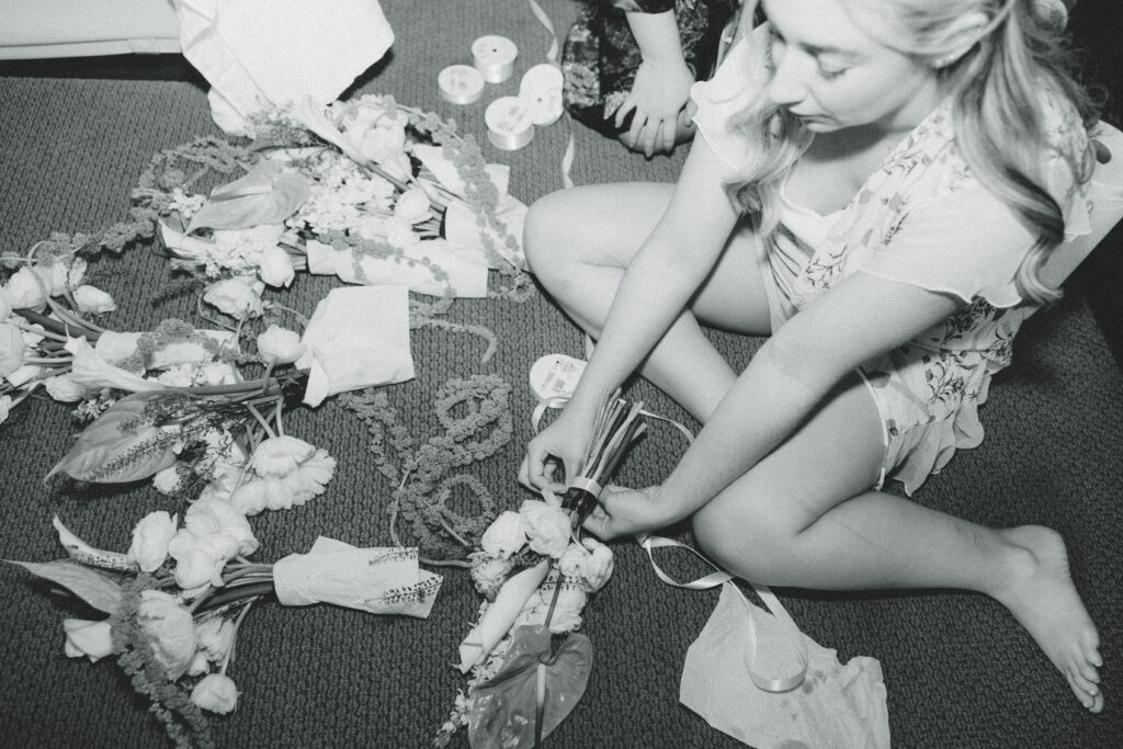 a bride sits on the hotel room floor and puts together her bridesmaids bouquet during her documentary style wedding photos