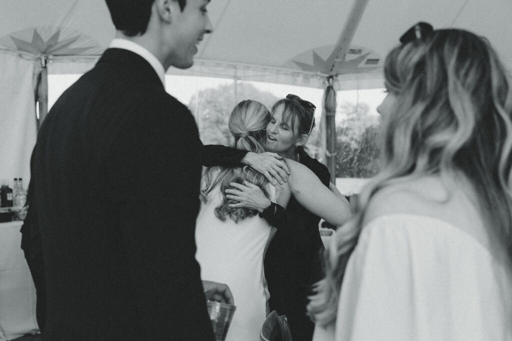 the bride shares a hug with a guest in the background as the groom speaks with another one during their reception with documentary style wedding photos