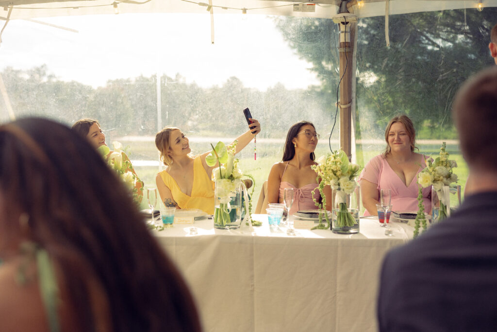 bridesmaids sit at the head table, one takes a selfie in documentary style wedding photos