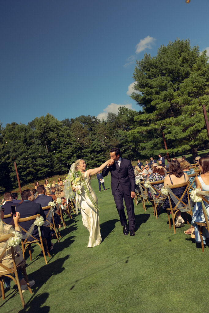 the bride reaches her arm out to her husband as they walk down the aisle together as he presses a kiss to her hand recreating an iconic JFK jr wedding photo in their documentary style wedding photos