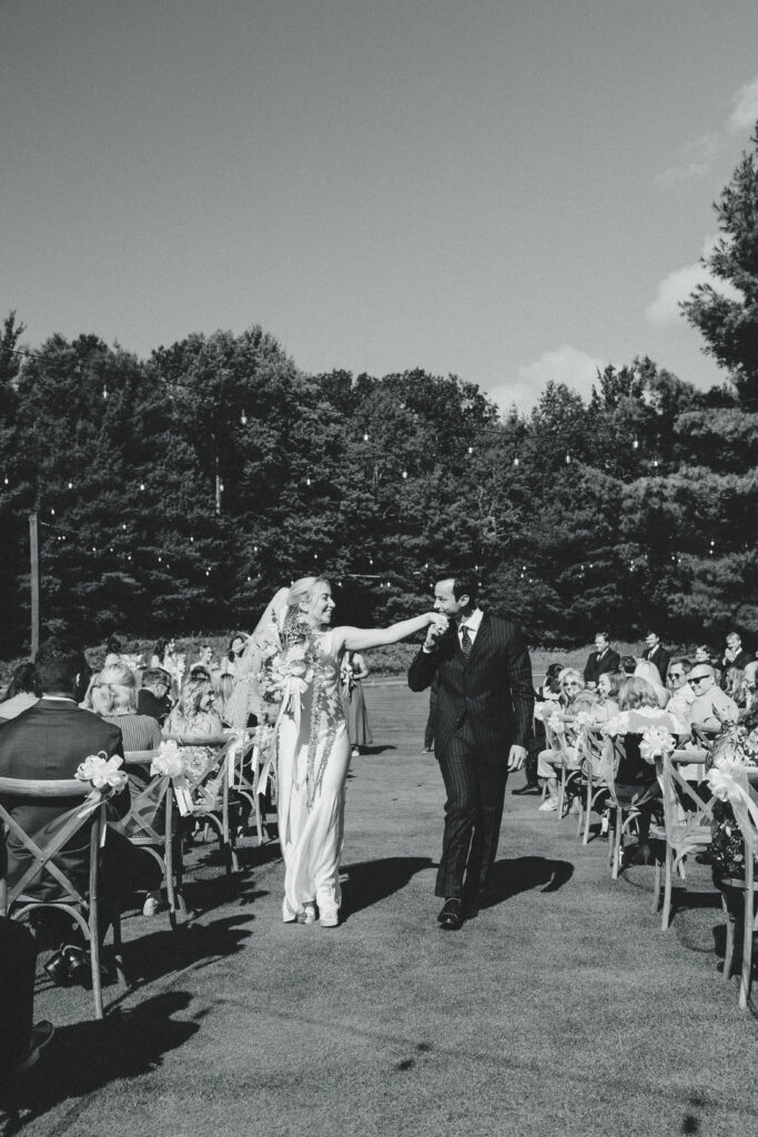 a bride and groom recreate jfk jr's wedding photo hand kiss while walking down the aisle together in this black and white documentary style wedding photos