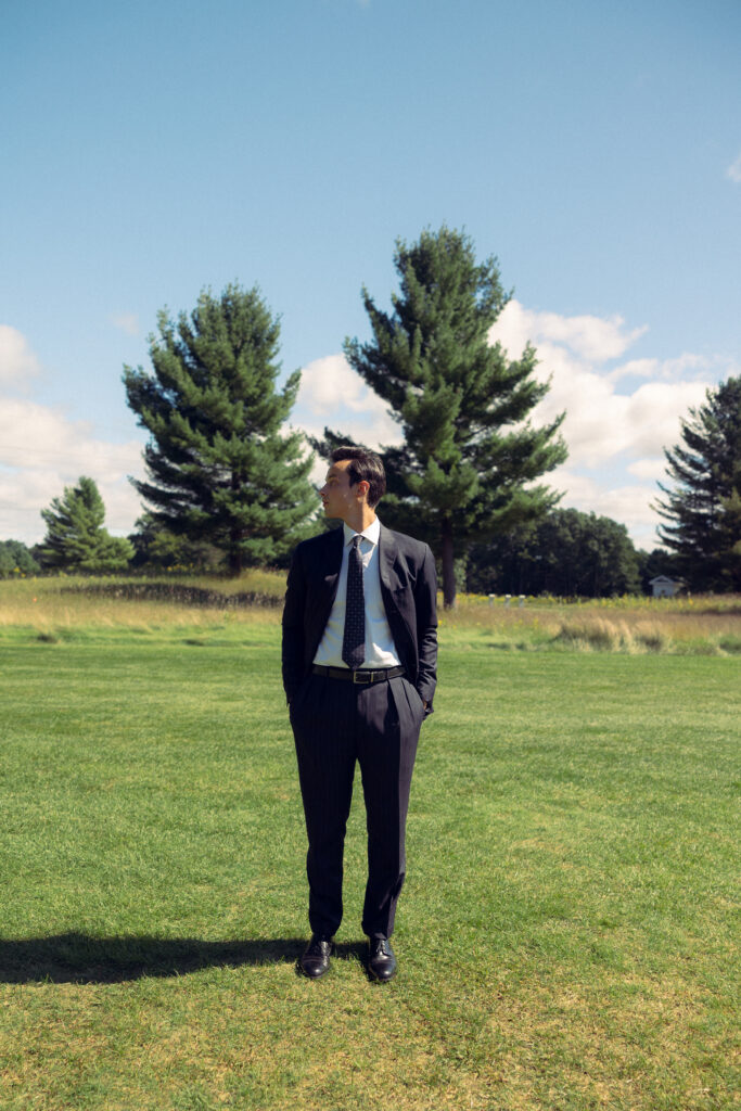 the groom stands in the grass looking to the side with his hands in his pocket with his shadow on the ground beside him