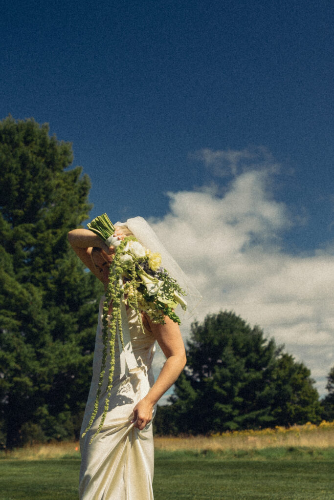 the bride shields her eyes from the sun with her bouquet in one hand and the other holding the length of her dress