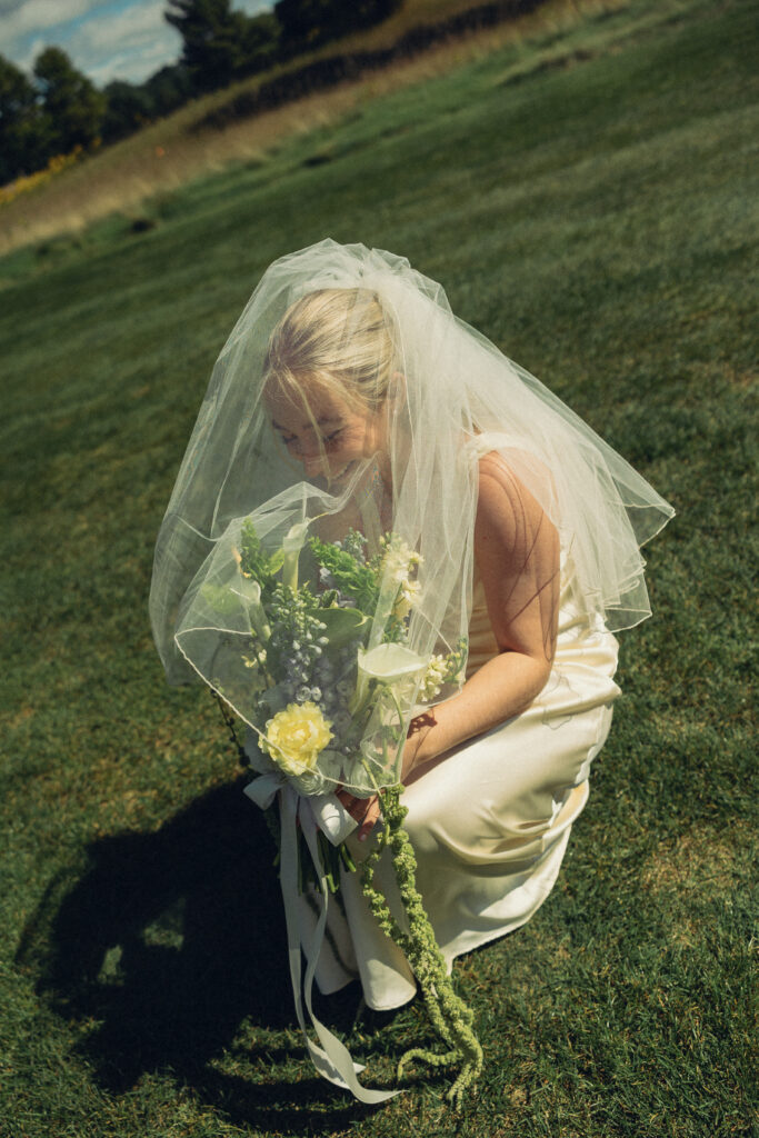 a bride laughs from under her veil as she crouches to the grass in documentary style wedding photos