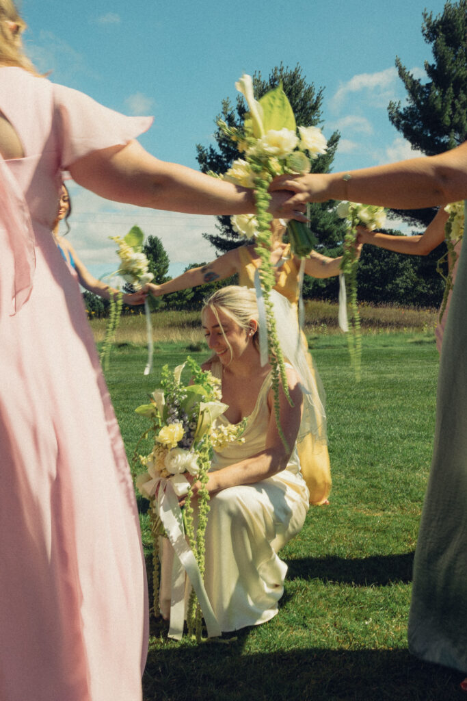 documentary style wedding photos where a bride squats down in her dress in the grass and smiles as the bridesmaids move in a circle around her