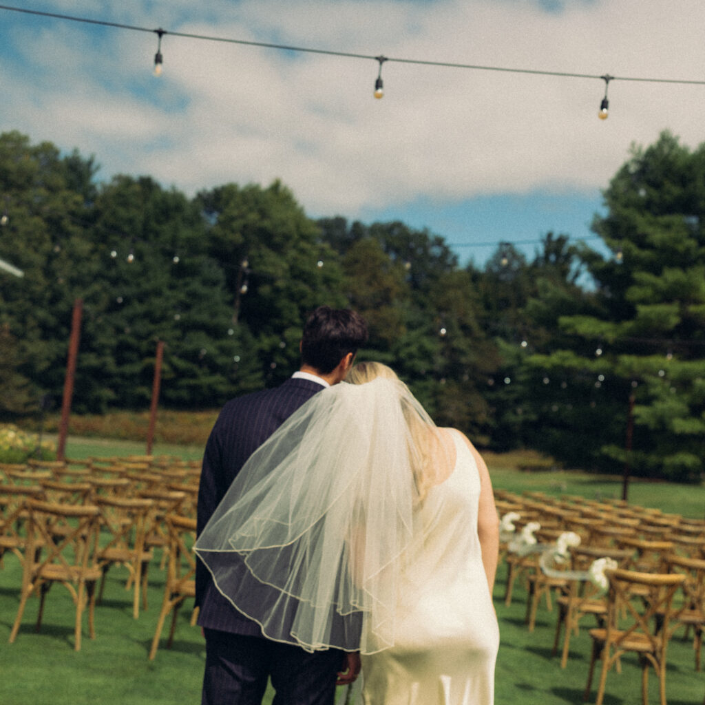 documentary style wedding photos a bride rests her head on her husband's shoulder as they look back at the site of their wedding ceremony