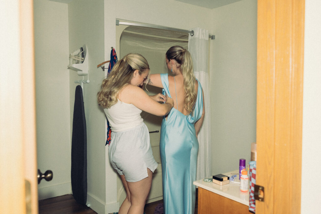 documentary style wedding photos, a bride helps her bridesmaid into her dress in the hotel bathroom in this view from the doorway