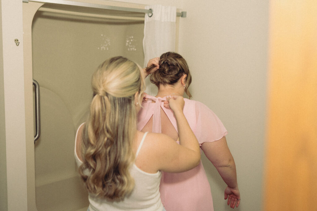 documentary style wedding photos a bride ties the bow of a bridesmaids dress in a hotel bathroom