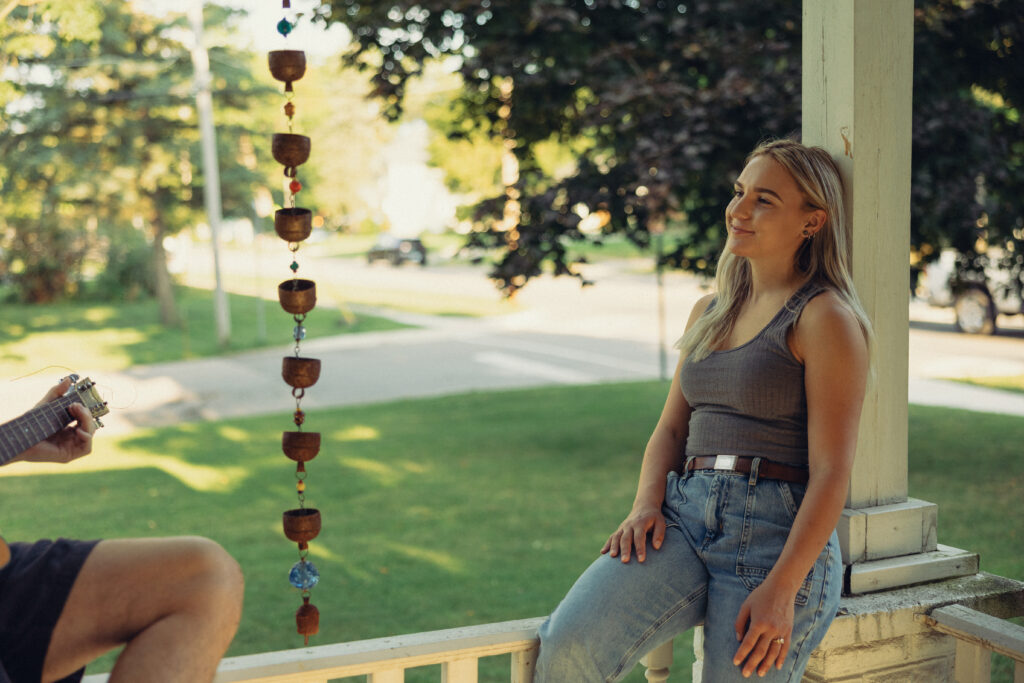 a woman leans against the post of her porch as her fiancee plays guitar for her in their 10 things i hate about you inspired engagement photos