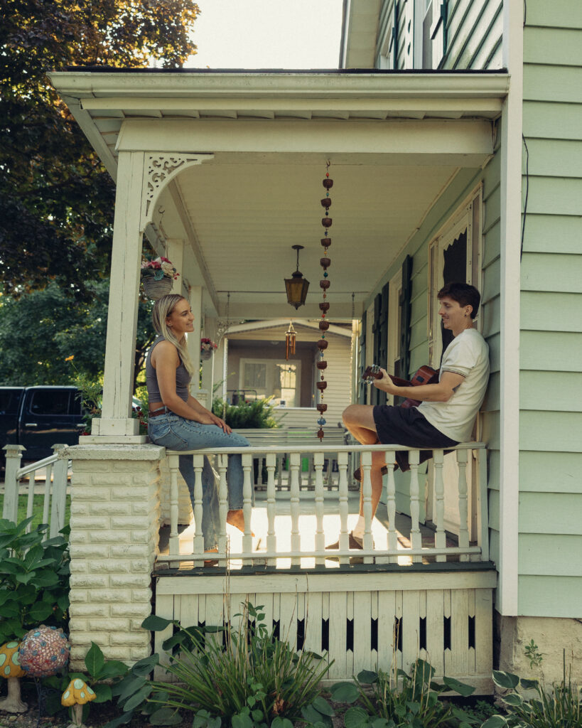 during their engagement photos a couple sits on the railing of their porch as he plays guitar for her in this cinematic feeling portrait