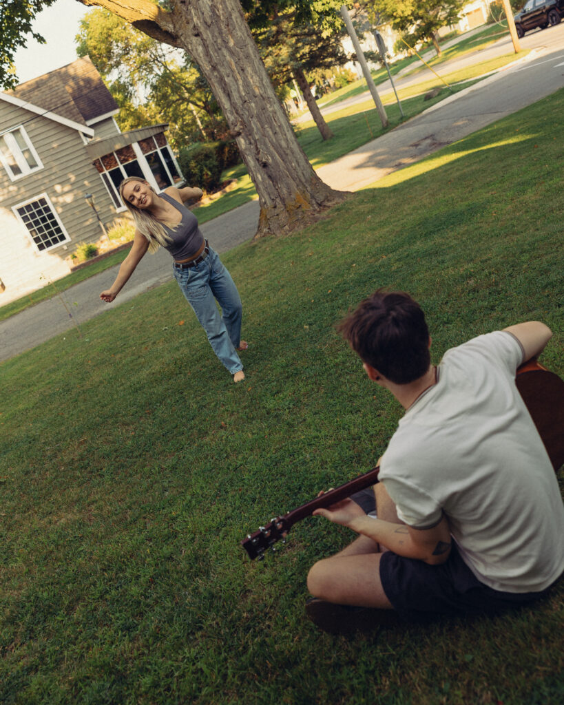 a woman dances in the yard as her fiancee sits playing guitar in front of her during their engagement photos