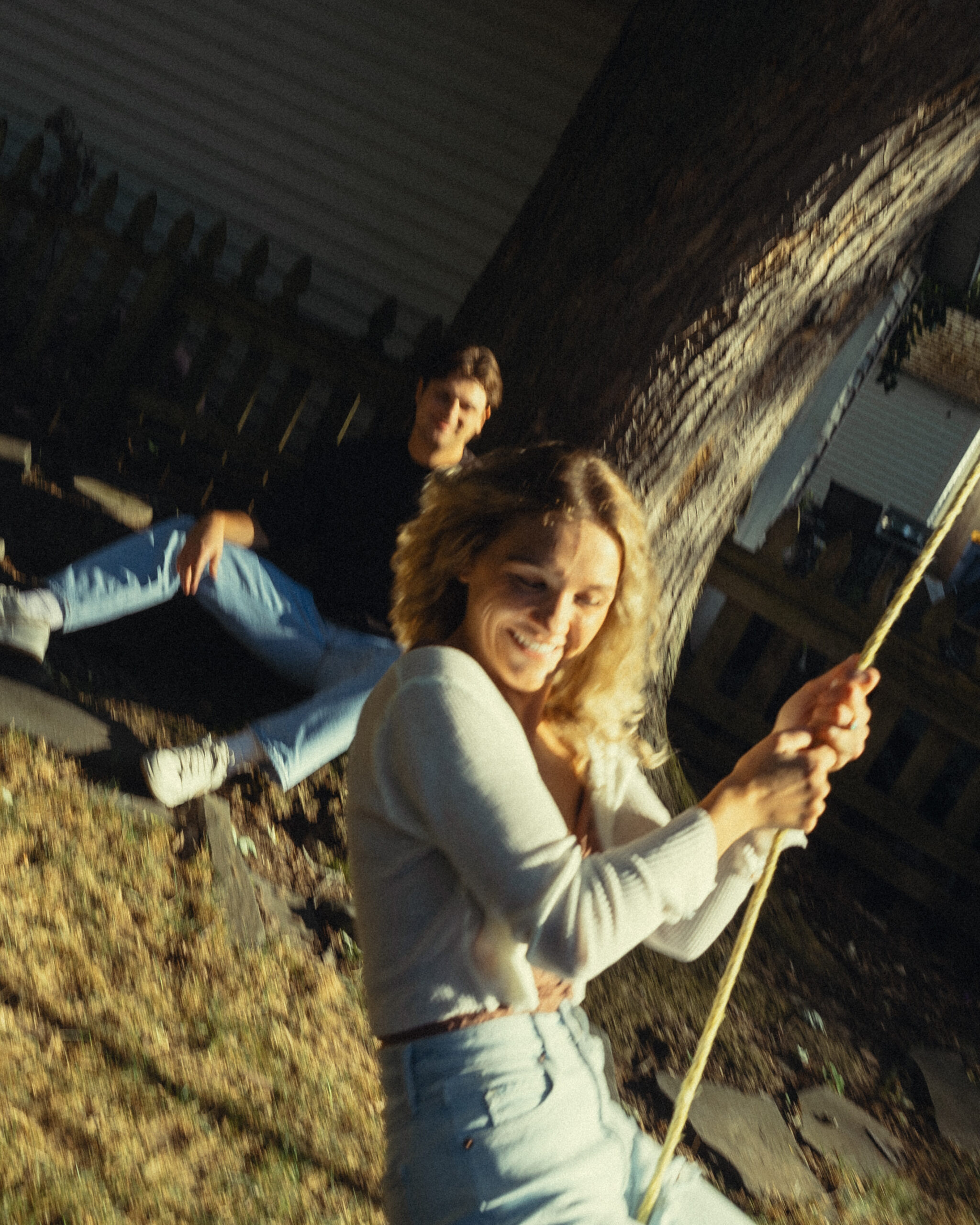 summer engagement photos where a woman smiles while swinging on a rope swing as her fiance smiles against a tree in the background