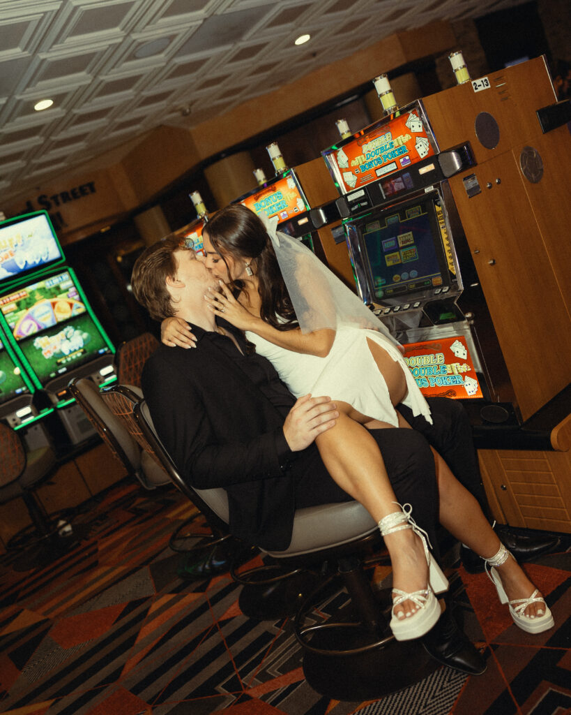 newlyweds share a kiss as she sits on his lap in front of a vegas casino machine during their vegas elopement photography
