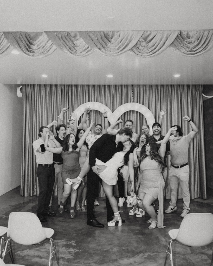 newlyweds kiss with a hand on her leg as she is dipped with their friends hyping them up behind them during their wedding photos in vegas