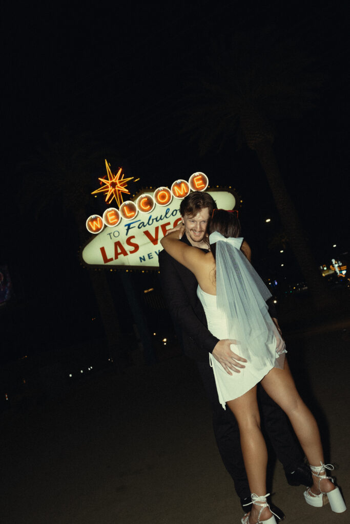 a man rests his hands on his wife's waist as she wraps her arms around his neck in front of the las vegas sign during their vegas elopement photography