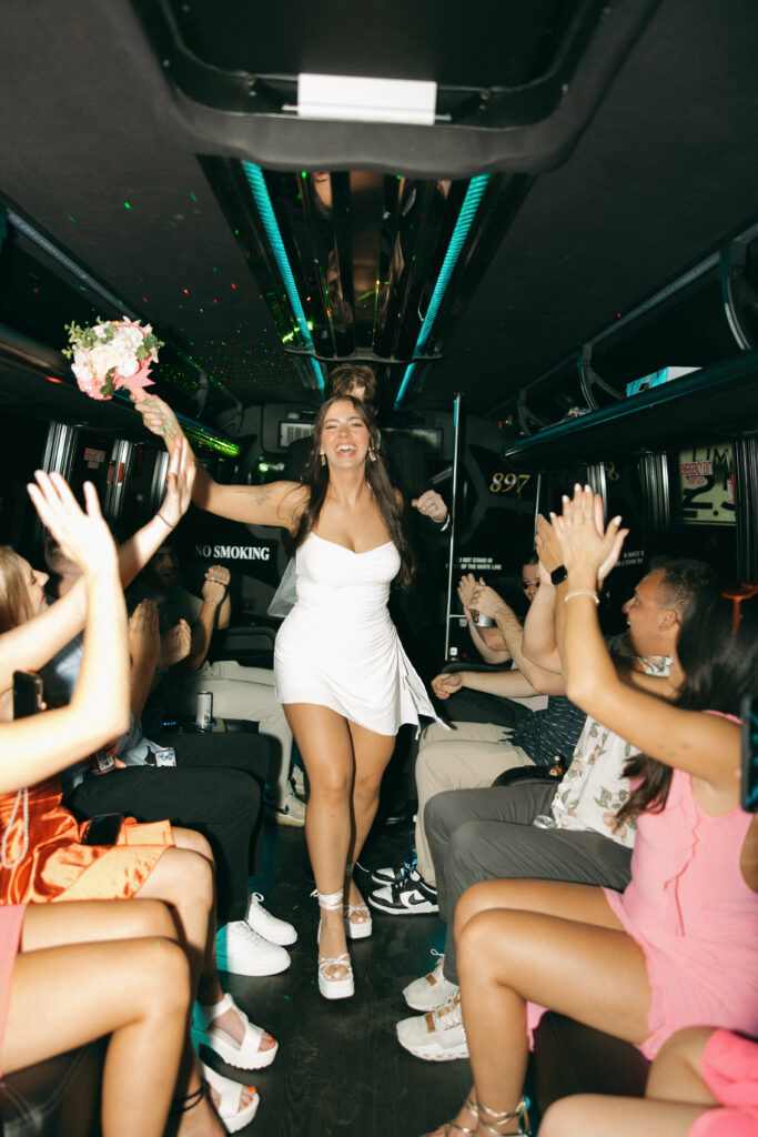 a smiling woman wearing a small white dress and holding a bouquet smiles as she boards a party bus during her vegas elopement photography