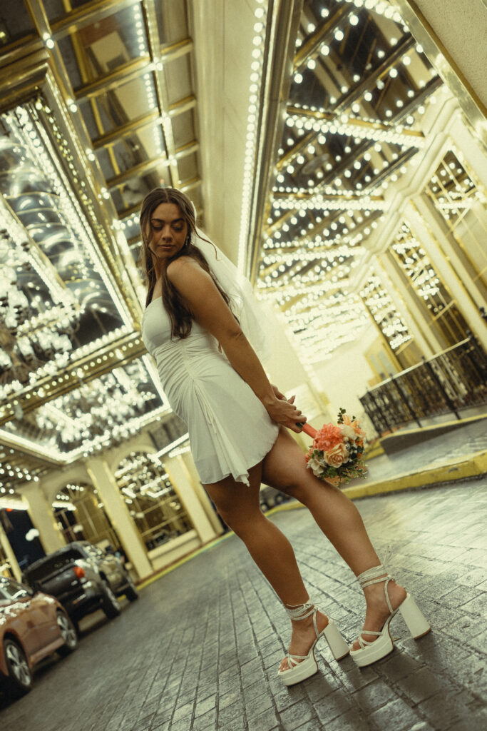 a woman wears a veil, a small white dress, and chunky white heels and holds a bouquet under sparkling vegas lights