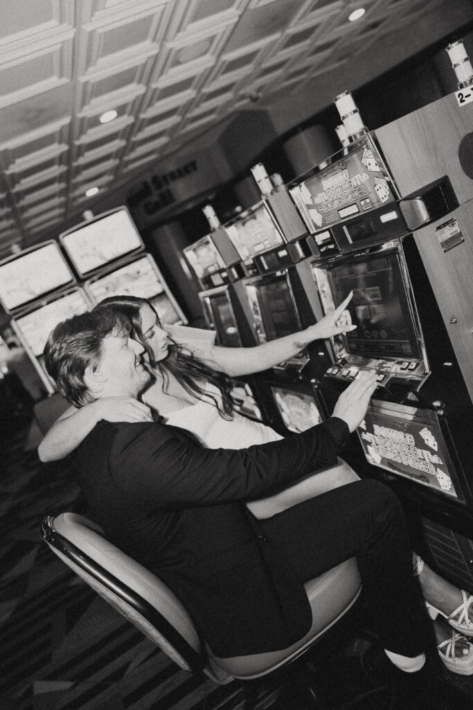 a woman sits on her husband's lap in front of a slot machine and points to a number during their vegas elopement photography