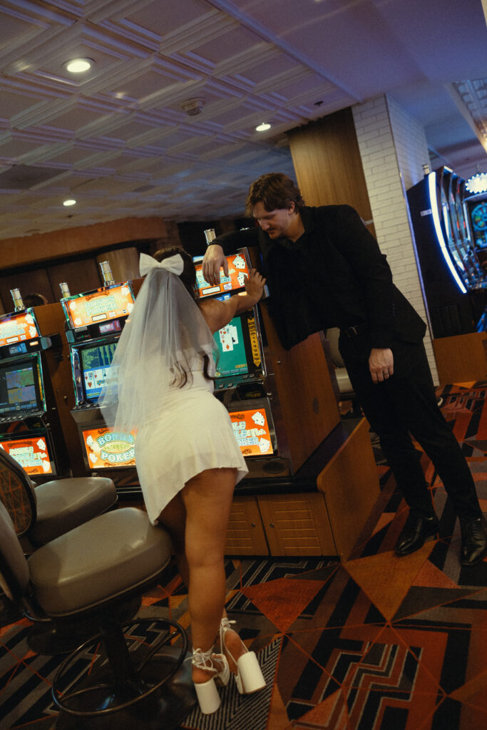 a woman leans against a poker simulation machine in a vegas casino next to her new husband during their wedding photos in vegas