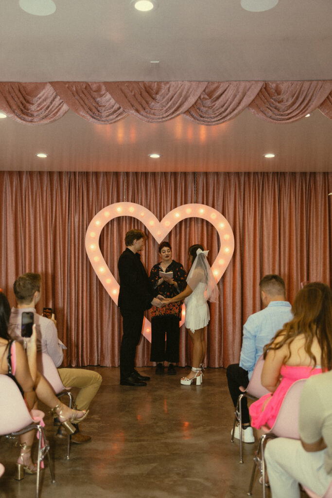 a couple stands in front of a heart shaped light holding each other's hands for their wedding photos in vegas