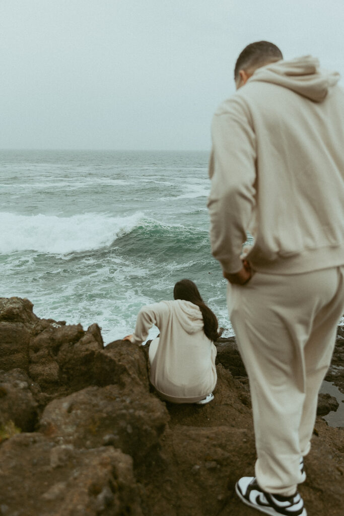 a woman crouches down on rocks in front of oregon waves as her husband stands behind her
