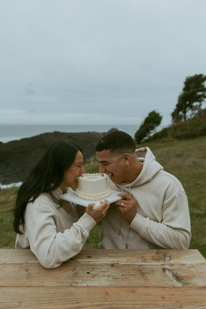 during their elopement photoshoot, a couple laughs as they take bites out of a heart shaped cake with the sea behind them