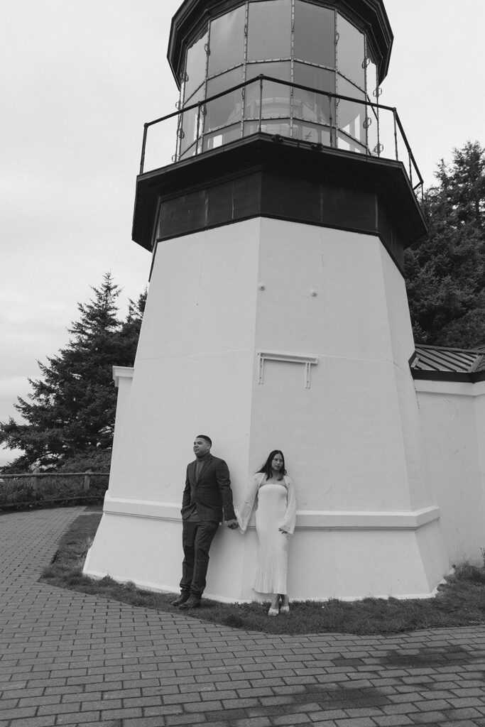 a black and white photo of a couple standing hand in hand while leaning against a lighthouse during their elopement photoshoot on the oregon coast