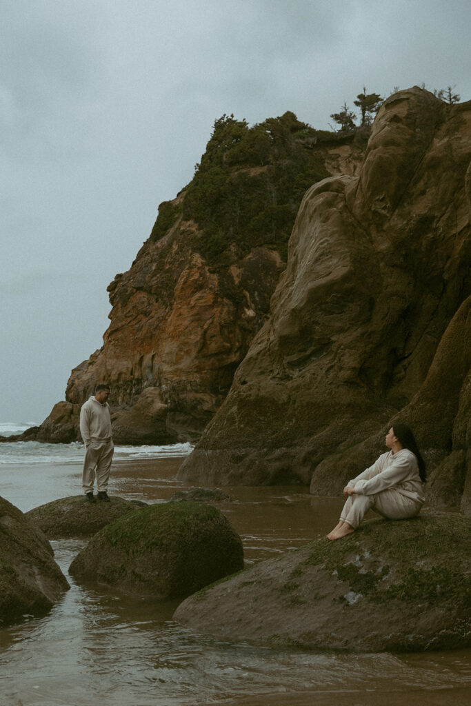 a woman sits on a rock and looks toward her husband that stands on another rock during their elopement photoshoot