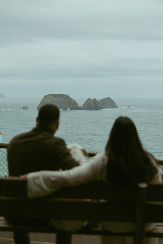 waves crashing over rocks are in focus framed by a couple sitting on the bench out of focus during their elopement photoshoot
