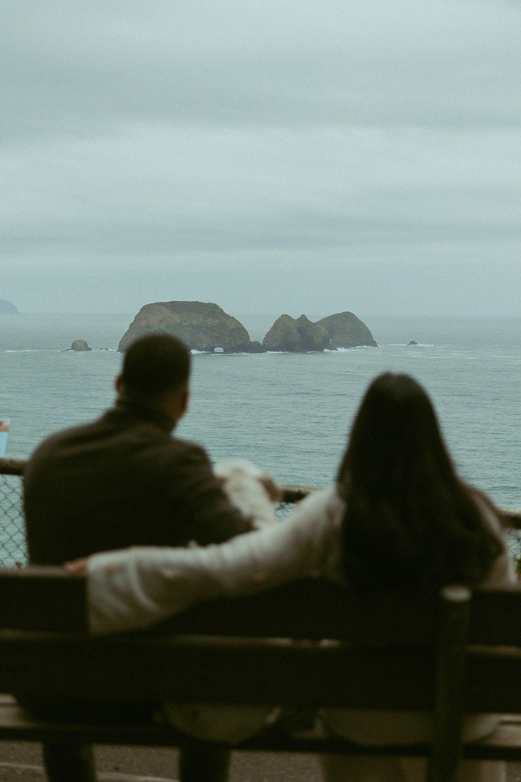 waves crashing over rocks are in focus framed by a couple sitting on the bench out of focus during their elopement photoshoot