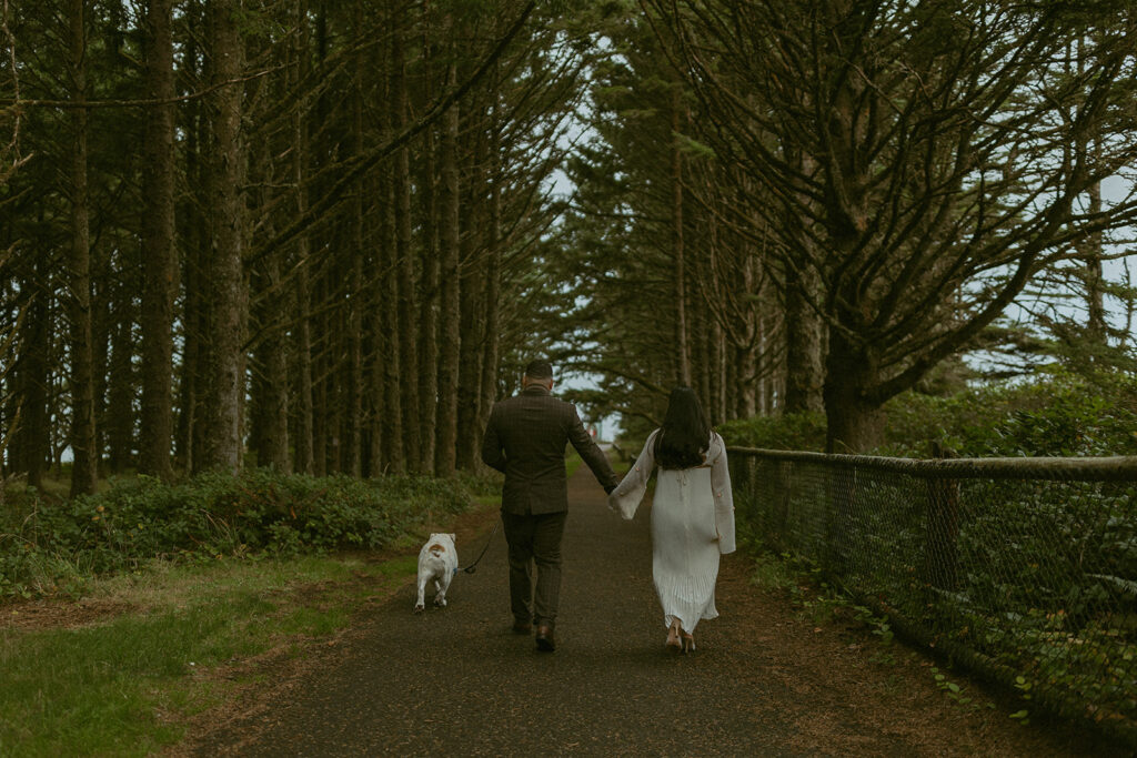 a couple walk on a paved path through a forest with their dog walking next to them during their elopement photoshoot
