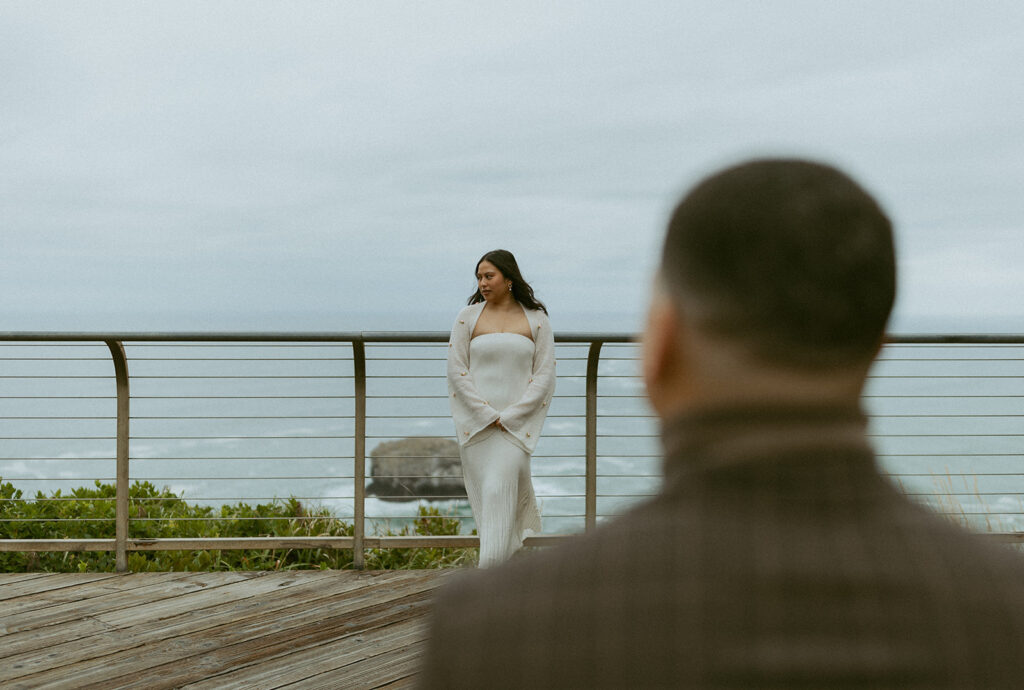 a woman stands against a railing on the edge of a cliff in front of the ocean while her husband who frames the shot out of focus faces her during their elopement photoshoot