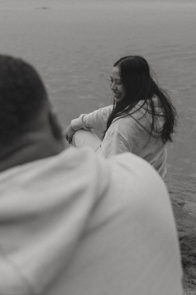 a woman smiles while sitting on a rock on the beach with her husband who faces her framing the shot during their elopement photoshoot