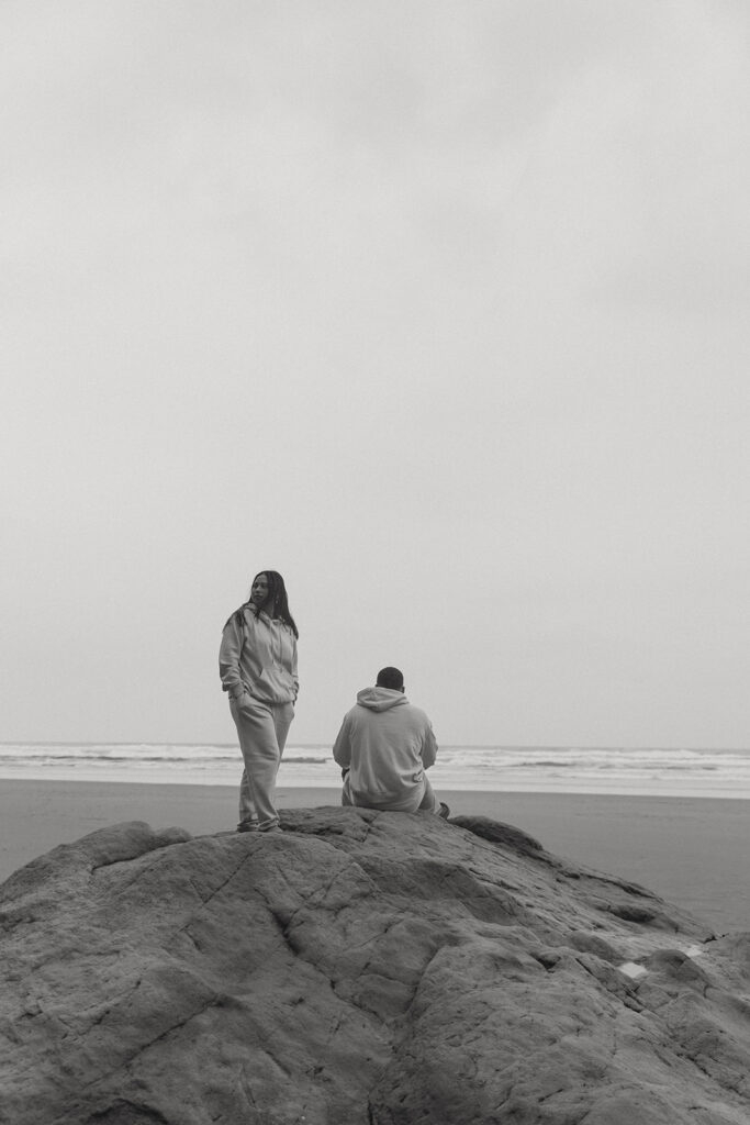 a woman stands on a rock next to her husband in front of the ocean during their elopement photoshoot