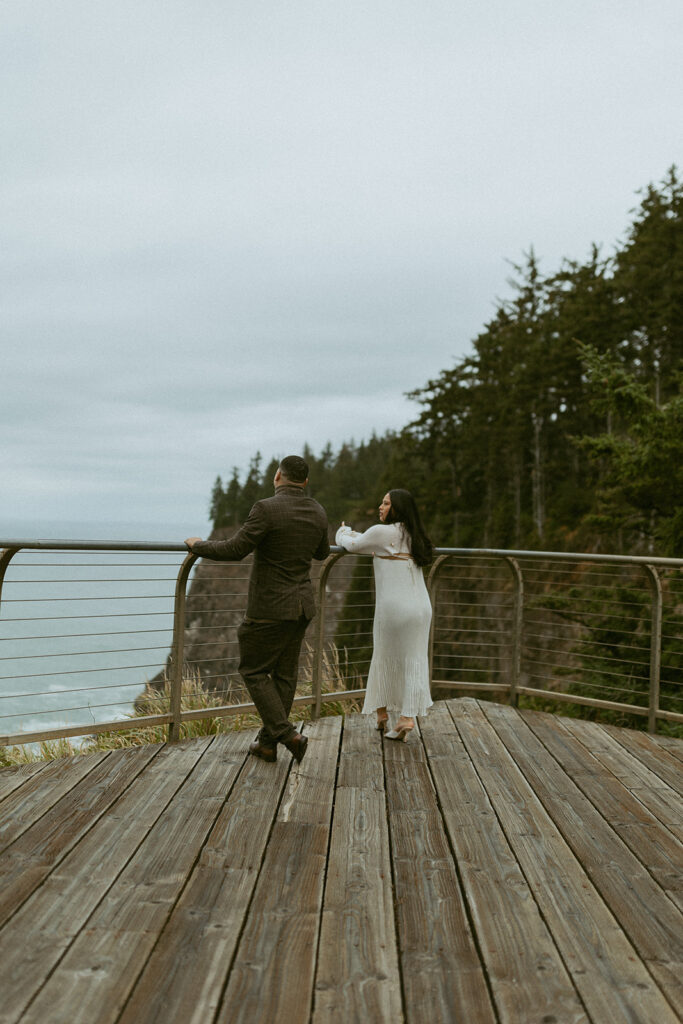 a couple stand a few feet apart on a lookout over the ocean leaning against a railing 