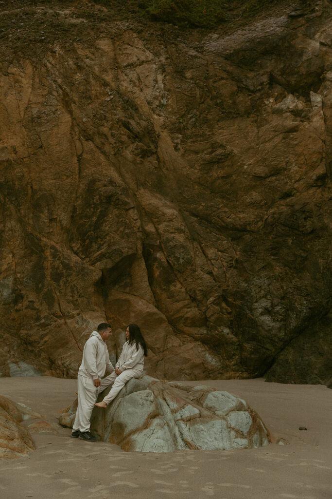 a woman sits on a rock facing her husband who stands at her eye level