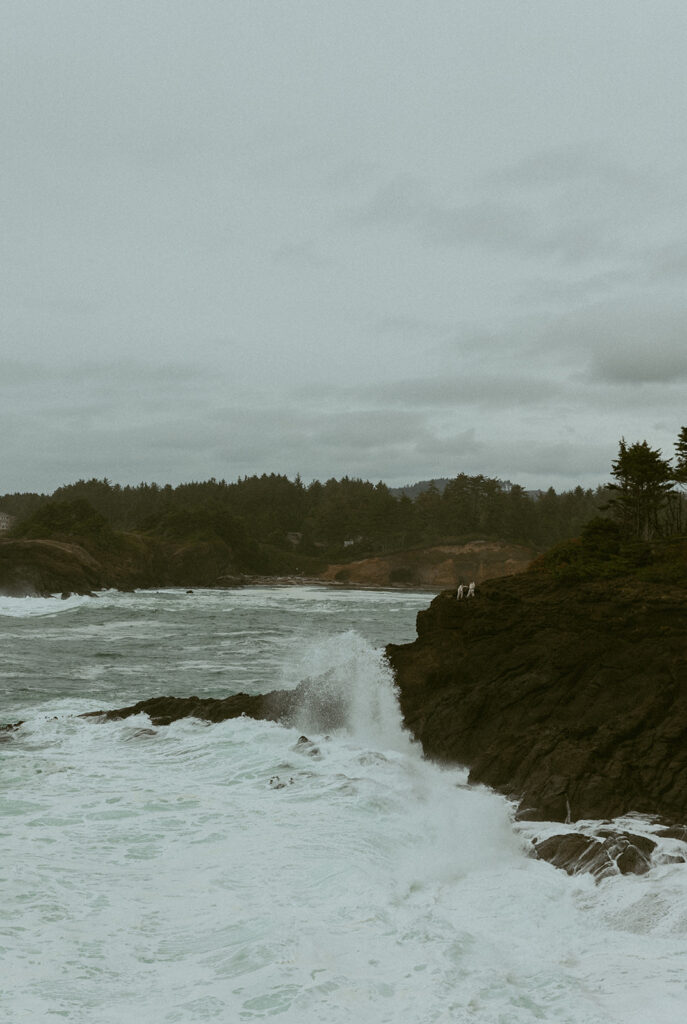 waves crash on rock formations on the oregon coast with two tiny figures standing far away in the distance from their oregon elopement photographer