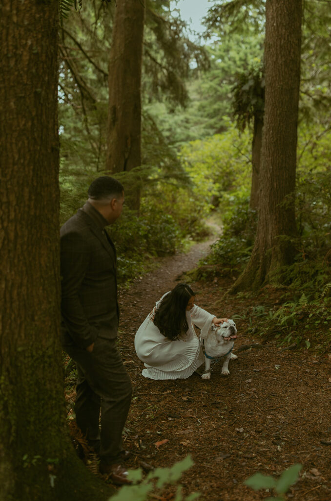 a woman crouches down to pet her dog as her husband looks toward her as they stand in the forest 