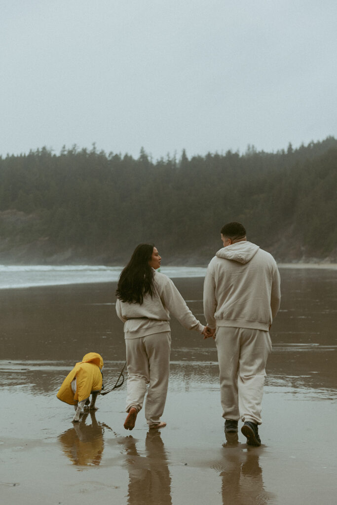 a couple wearing matching sweatsuits walk on the beach with their dog that wears a yellow rain jacket during a shoot with their oregon elopement photographer