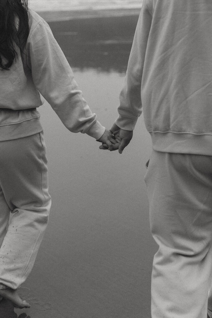 a couple holds hands on the beach on a rainy day wearing matching sweatsuits in this cropped photo from their elopement photoshoot