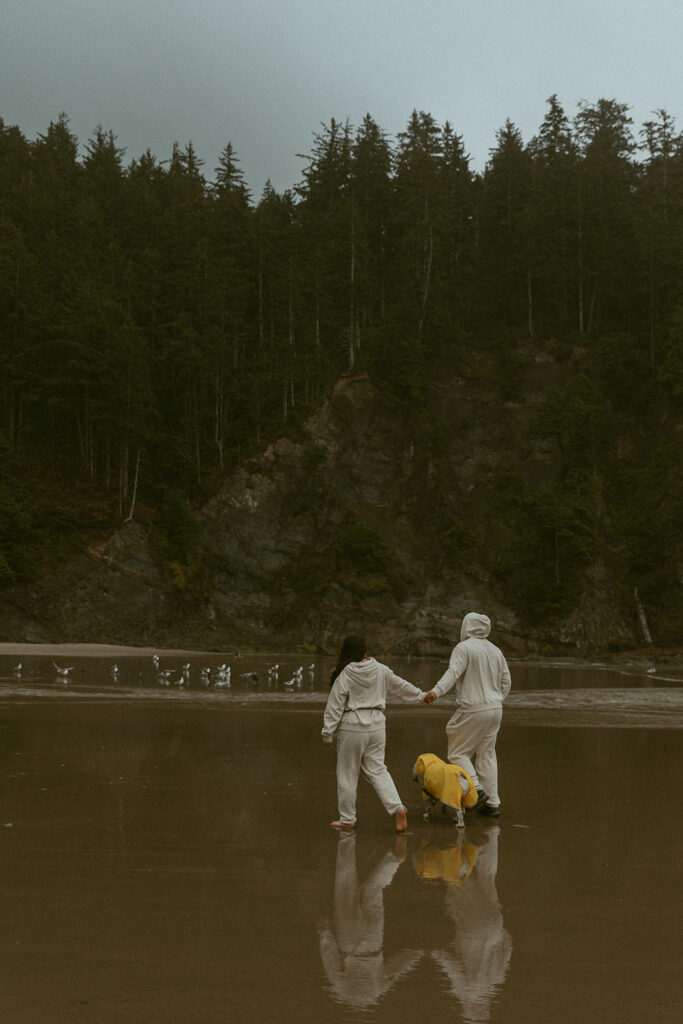 a couple holds hands wearing matching sweatsuits on the oregon coast with their dog, wearing a yellow rain jacket, who walks between them