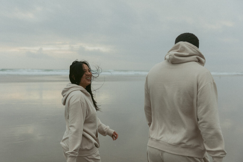 a woman smiles while walking on the beach with her husband in matching sweatsuits during their elopement photoshoot