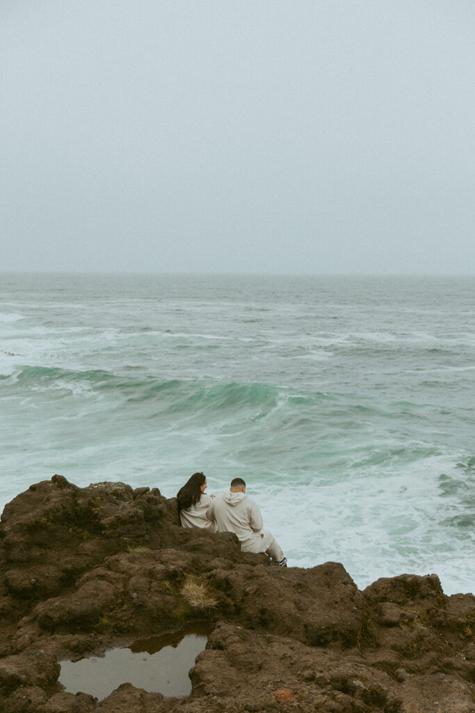 a couple sits together on the rocks in front of the ocean