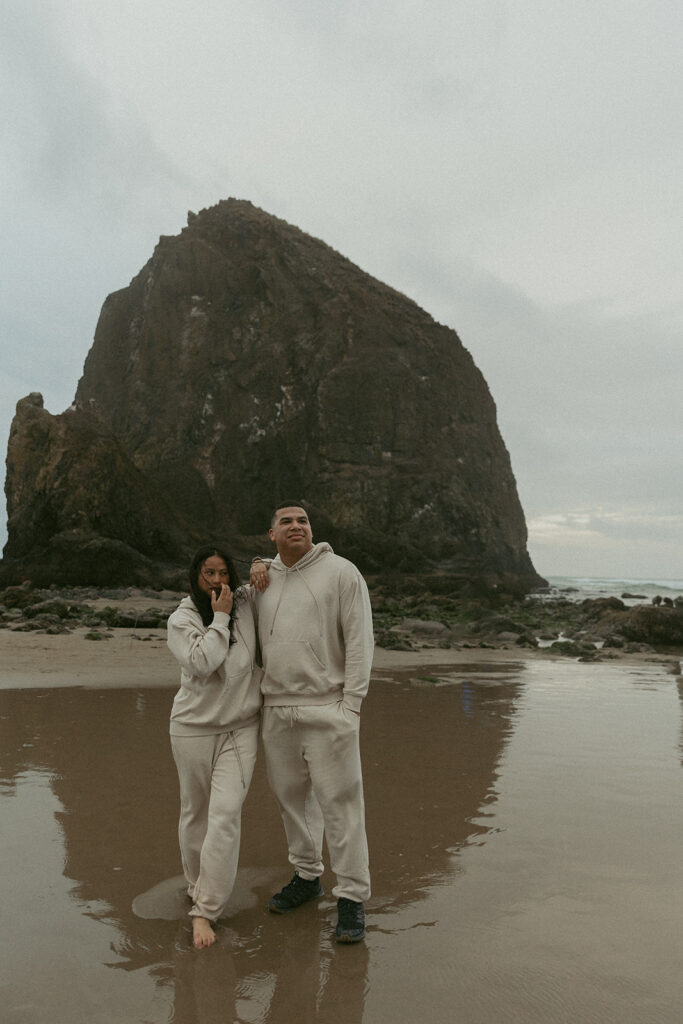 a woman touches her face while resting an arm on her husband's shoulder as they stand on the beach in matching sweatsuits during their elopement photoshoot