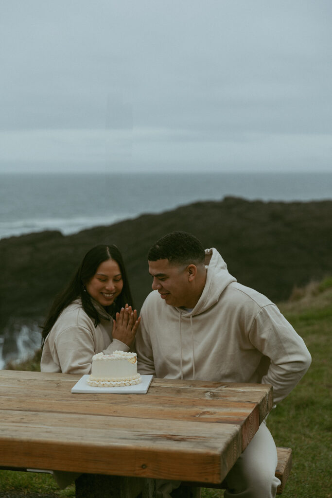 during their elopement photoshoot, a couple sits at a picnic bench in front of the seaside looking down at a heart shaped cake