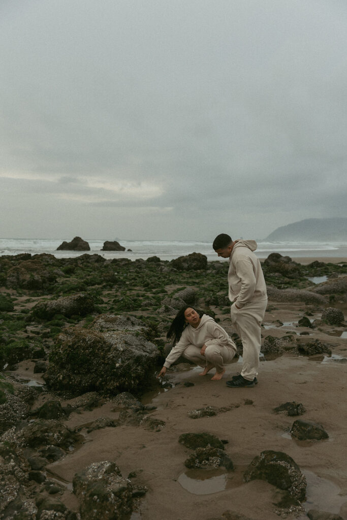 a couple explores the rocky beach in front of oregon waves during, she crouches down to point at something as her husband looks down at her